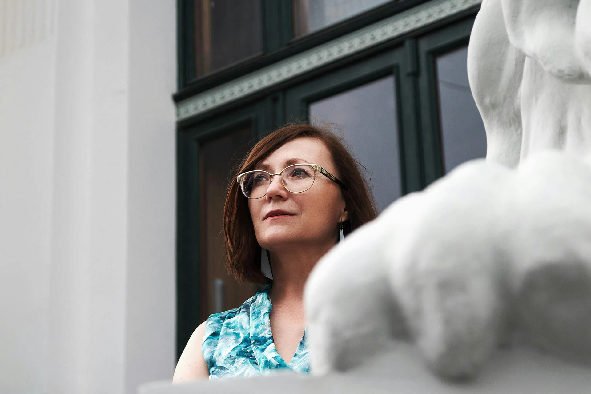 A middle-aged woman with glasses gazing off into the distance while standing on a balcony.